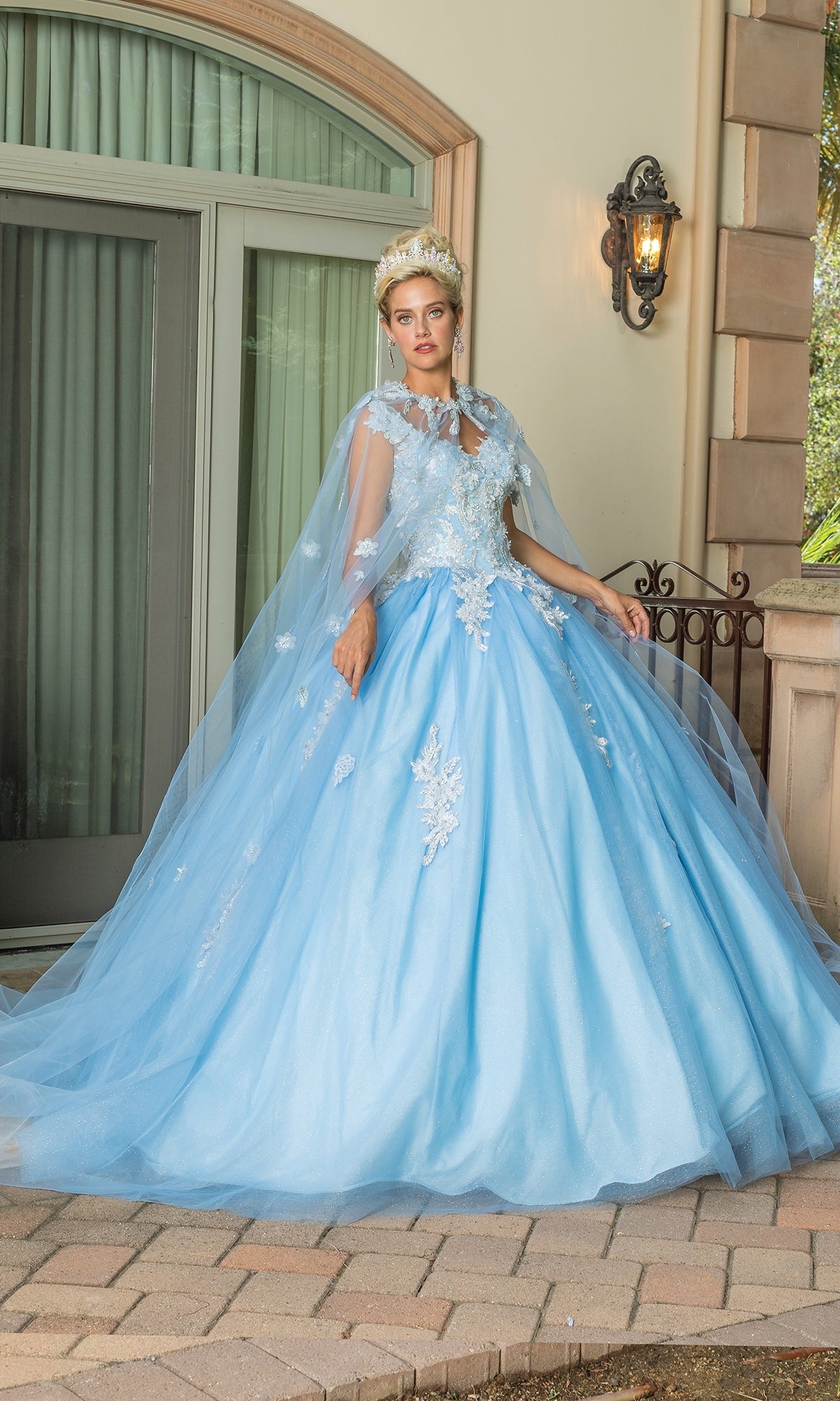 A woman in the Quinceanera Dress 1597 by Dancing Queen—light blue with white floral appliqué, cap sleeves, and a matching cape—stands on a stone path outside a building with a glass door and lantern.